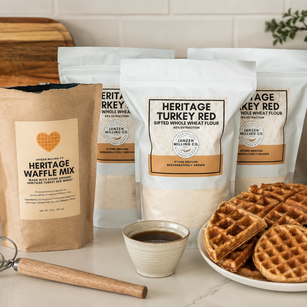 Packages of Janzen Milling Co. flour and waffle mix with a plate of waffles and a cup of coffee on a kitchen counter.